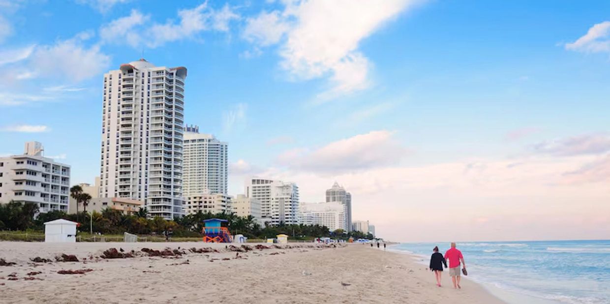 Couple walking along a sandy beach with city buildings in the background.