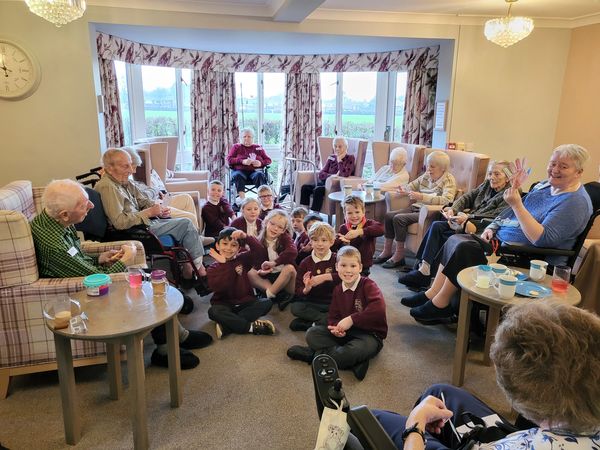 Children and elderly people gathered in a cozy room, smiling and waving.