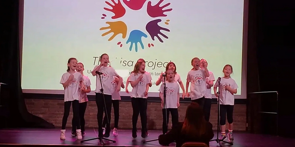 Children singing on stage wearing matching T-shirts with a colorful hand logo projected behind.