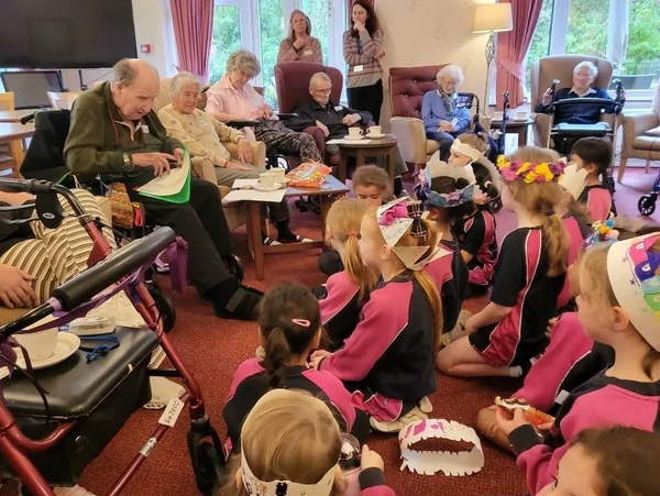 Children in colorful hats sitting on the floor, engaged with elderly people in a cozy room.
