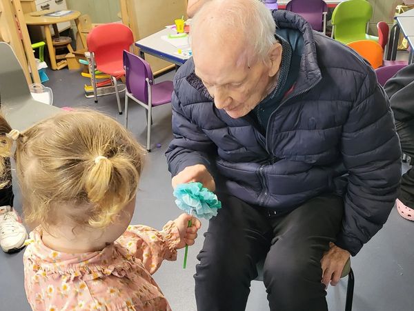 A young girl hands a blue flower to an elderly man in a cozy room.
