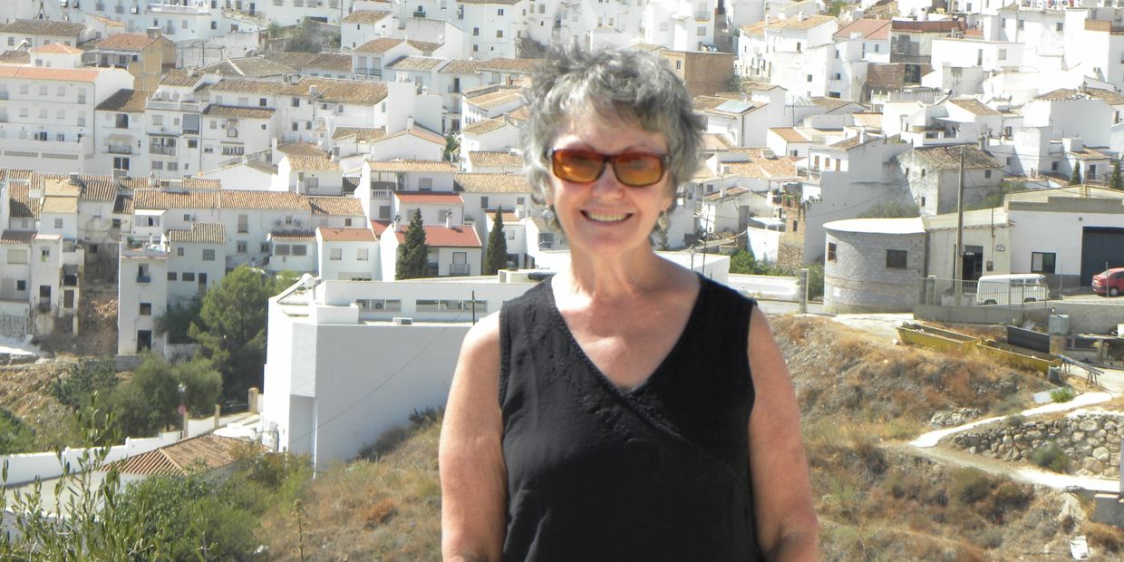 Smiling woman in sunglasses stands against a backdrop of white village houses on a sunny day.