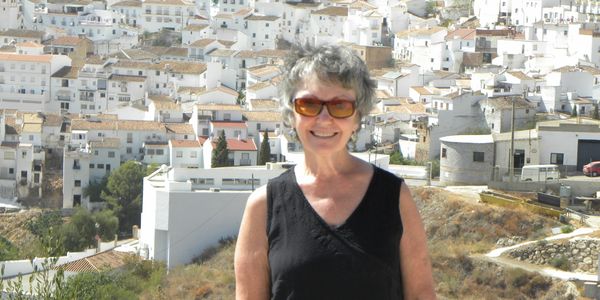 Smiling woman wearing sunglasses stands in front of a hillside town with white buildings.
