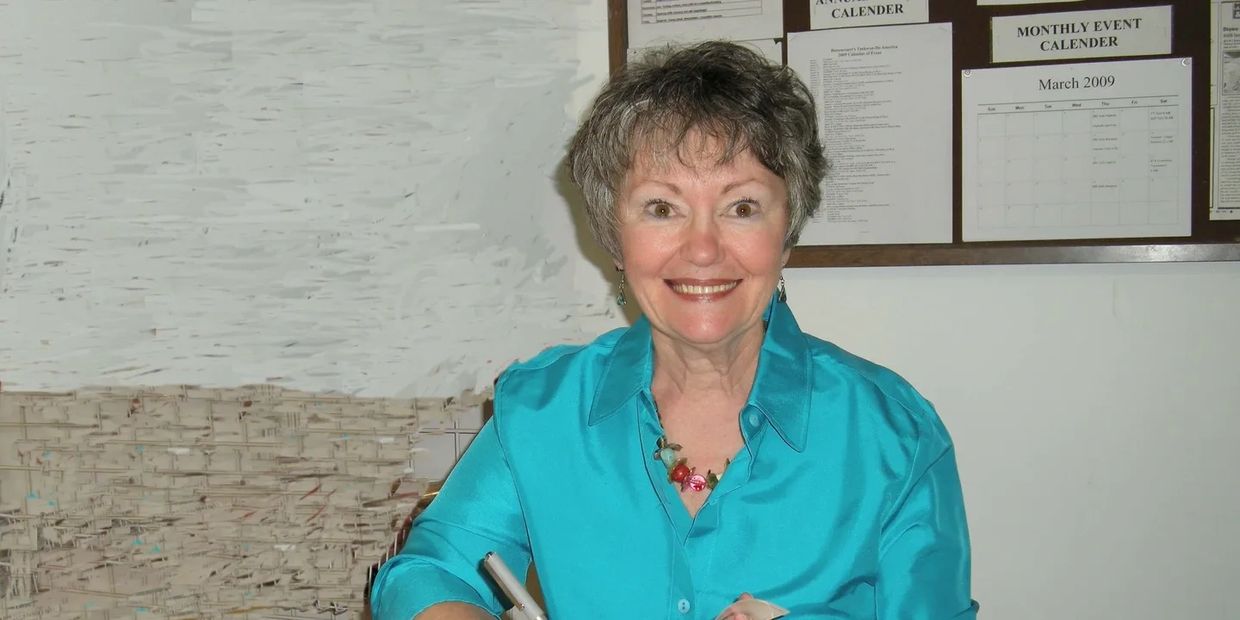 Smiling older woman in a teal shirt signs a book indoors.
