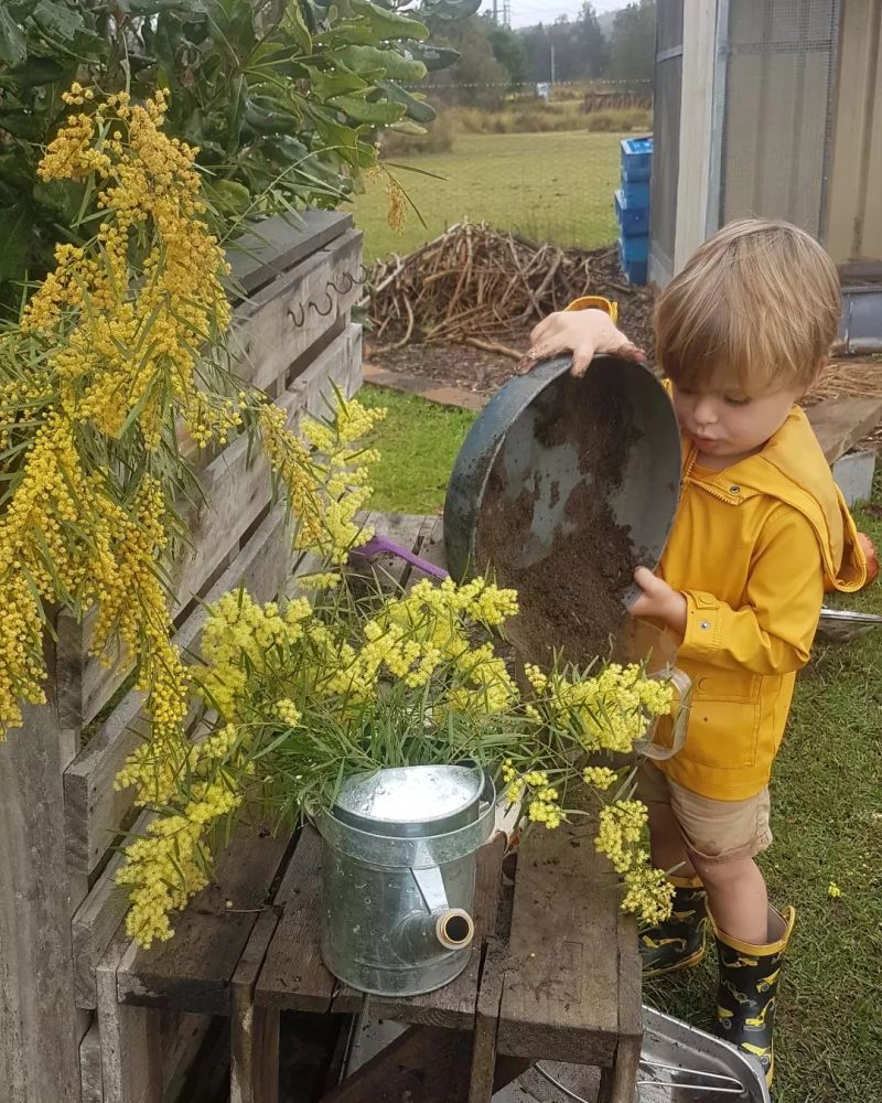 Child in yellow raincoat pouring soil near yellow flowers and watering can.