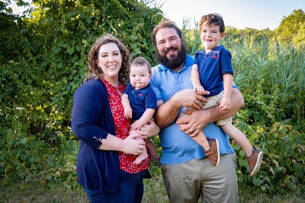Happy family of four posing outdoors in a green, sunny setting.