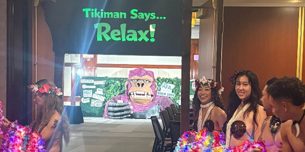 A group of Polynesian performers stand in two lines inside a hotel or event hallway, holding bright,