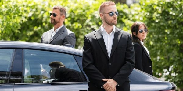 Three bodyguards in black suits and sunglasses stand alert near a black car.