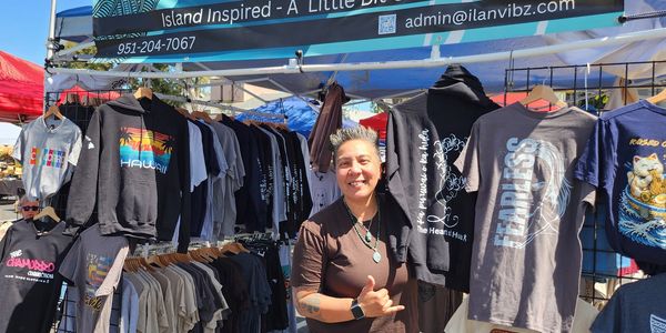 Vendor smiling in front of island-inspired clothing at outdoor market booth.