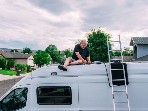 Man sitting on the roof of a white van working, with a ladder leaned against the side.