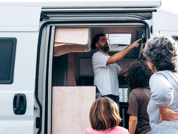 Man standing in the open side door of a van, pointing to the interior for a family looking on.