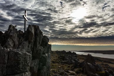 A cross stands on rugged rocks under a dramatic cloudy sky at sunset.