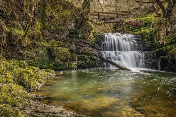 A serene waterfall with a wooden bridge above and moss-covered rocks.