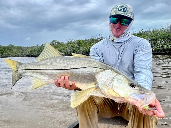 A smiling man holding a large fish while sitting in a boat on a cloudy day.