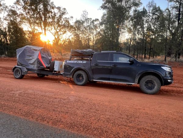 Black pickup truck towing covered equipment on a red dirt road at sunset.