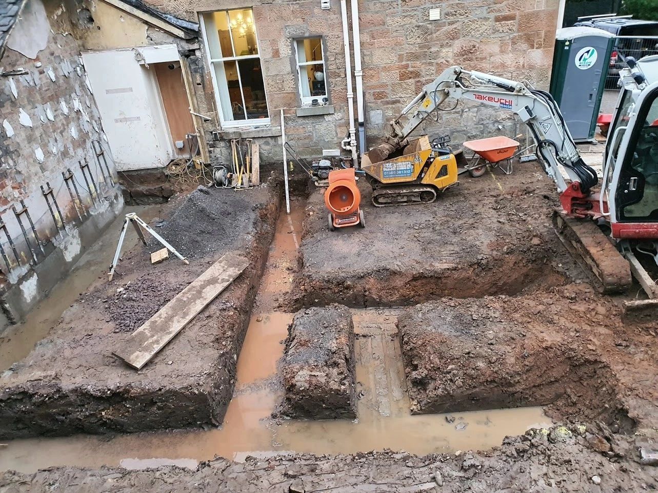 Excavation site beside a stone building with construction equipment and muddy trenches.