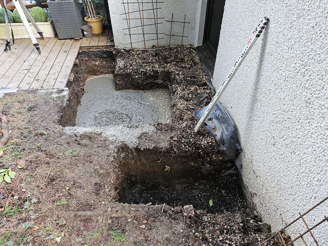 Construction site with dug soil, wet concrete, and measurement rod against a wall.