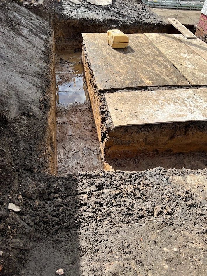 Construction site with a muddy trench and wooden boards covering an excavation.