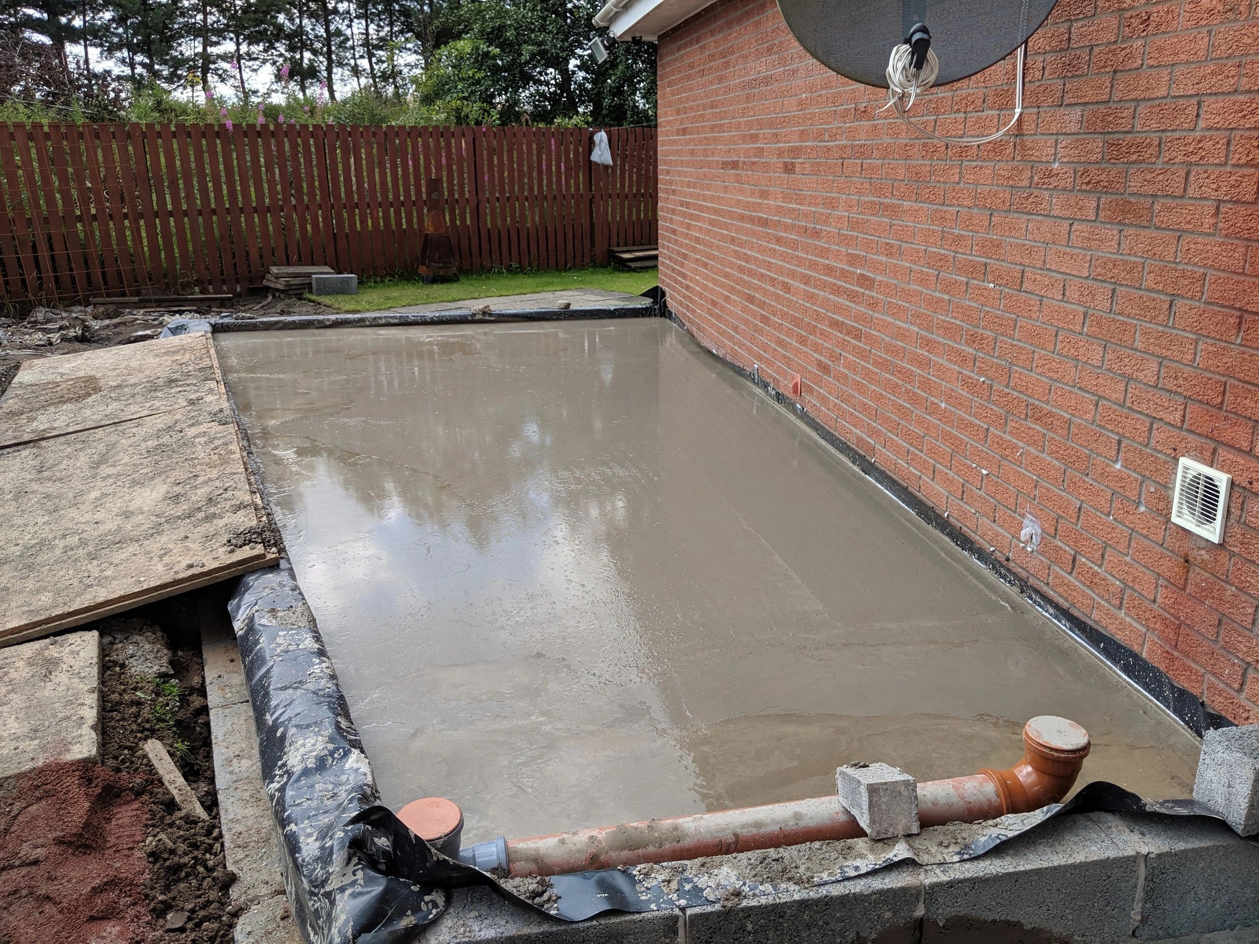 Freshly poured concrete slab next to a brick house with visible plumbing.