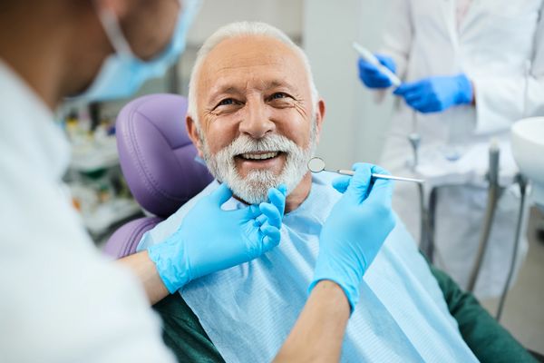 A smiling elderly man with a white beard is seated in a dental chair. 