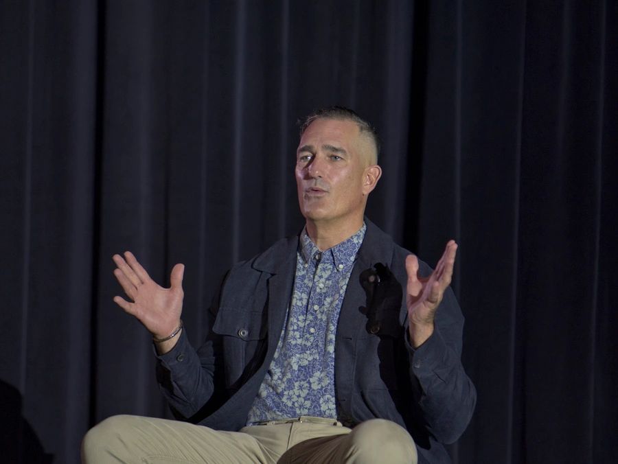 Man in patterned shirt and jacket speaking with expressive hand gestures.