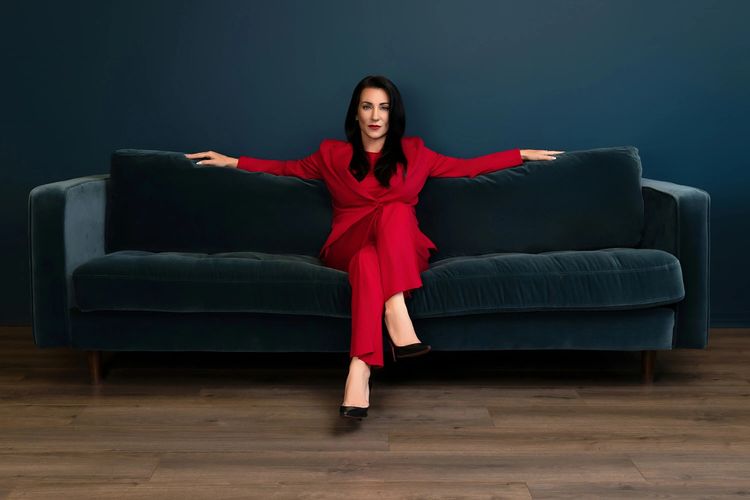 Dayna in red suit sitting confidently on dark sofa.