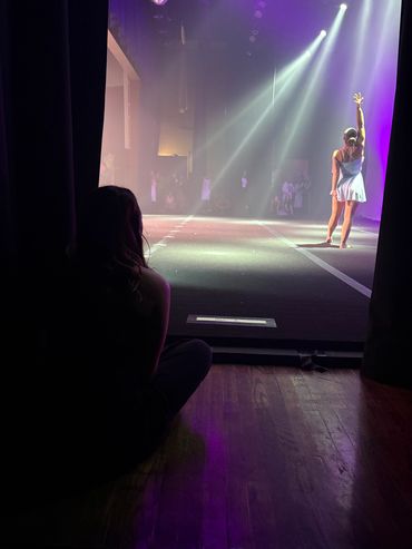 A dancer performs on stage as another watches from backstage.