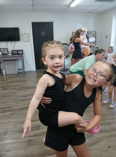 Two young girls in a dance studio, one holding the other and smiling.