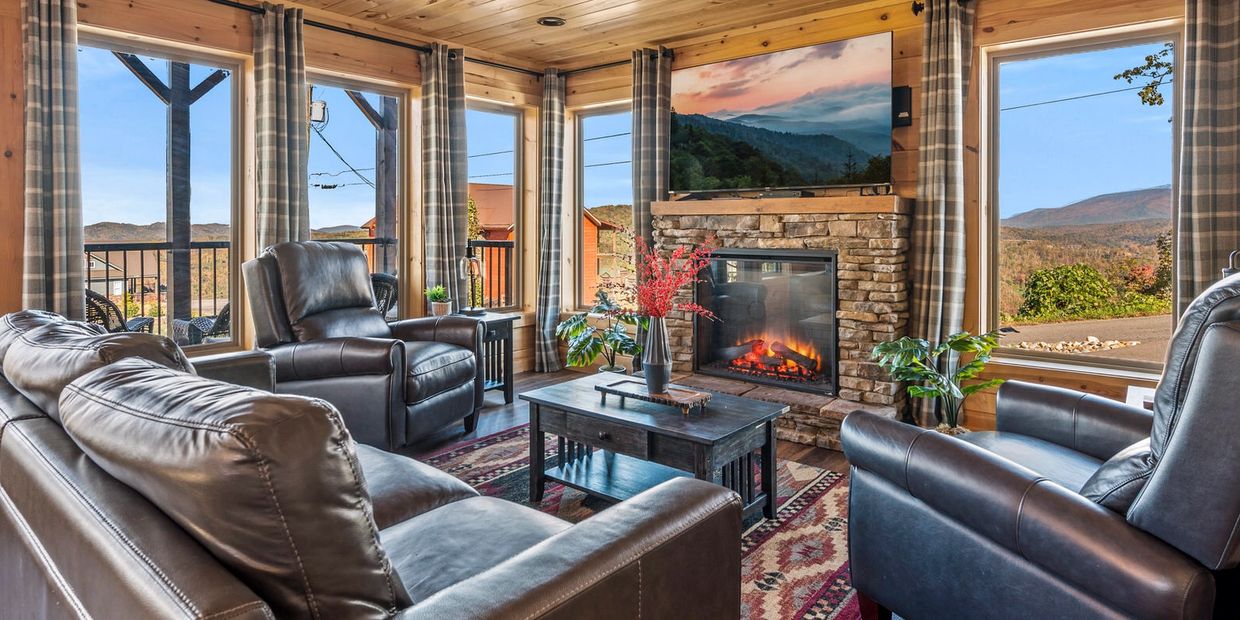 Cozy living room with leather furniture, stone fireplace, and large windows showing mountain views.