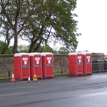 Red portable toilets lined up outdoors near a stone wall and trees.