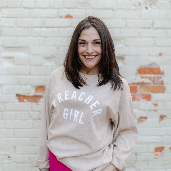 Smiling woman wearing a "Preacher Girl" sweatshirt stands against a brick wall.