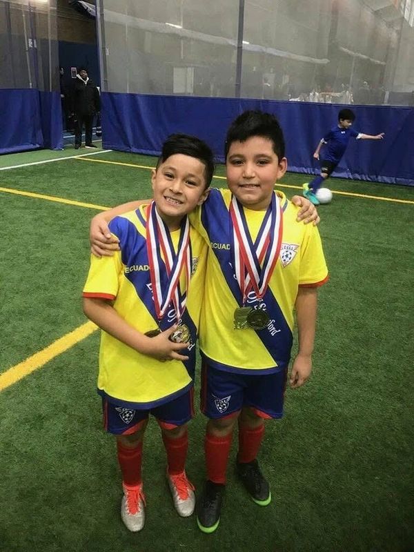 Two boys in soccer uniforms proudly wearing medals on an indoor field.