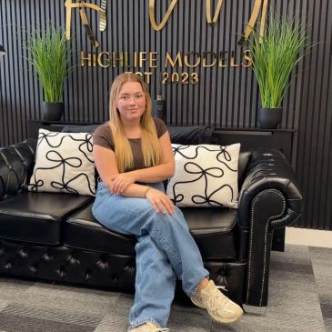 Young woman sitting on a black leather couch in front of a modeling agency sign.