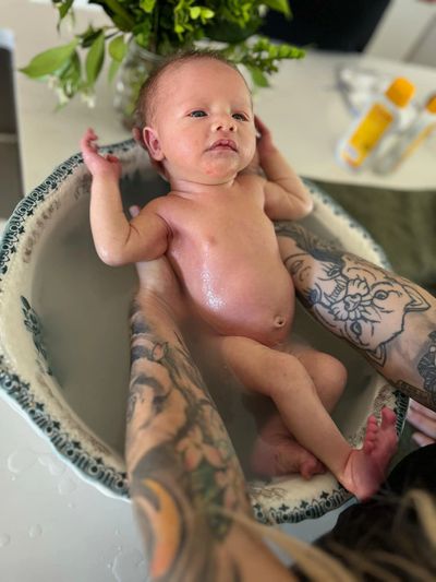 Newborn baby being bathed in a small tub, held by tattooed arms.