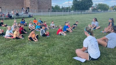 Children seated on grass listening to instructors outdoors.