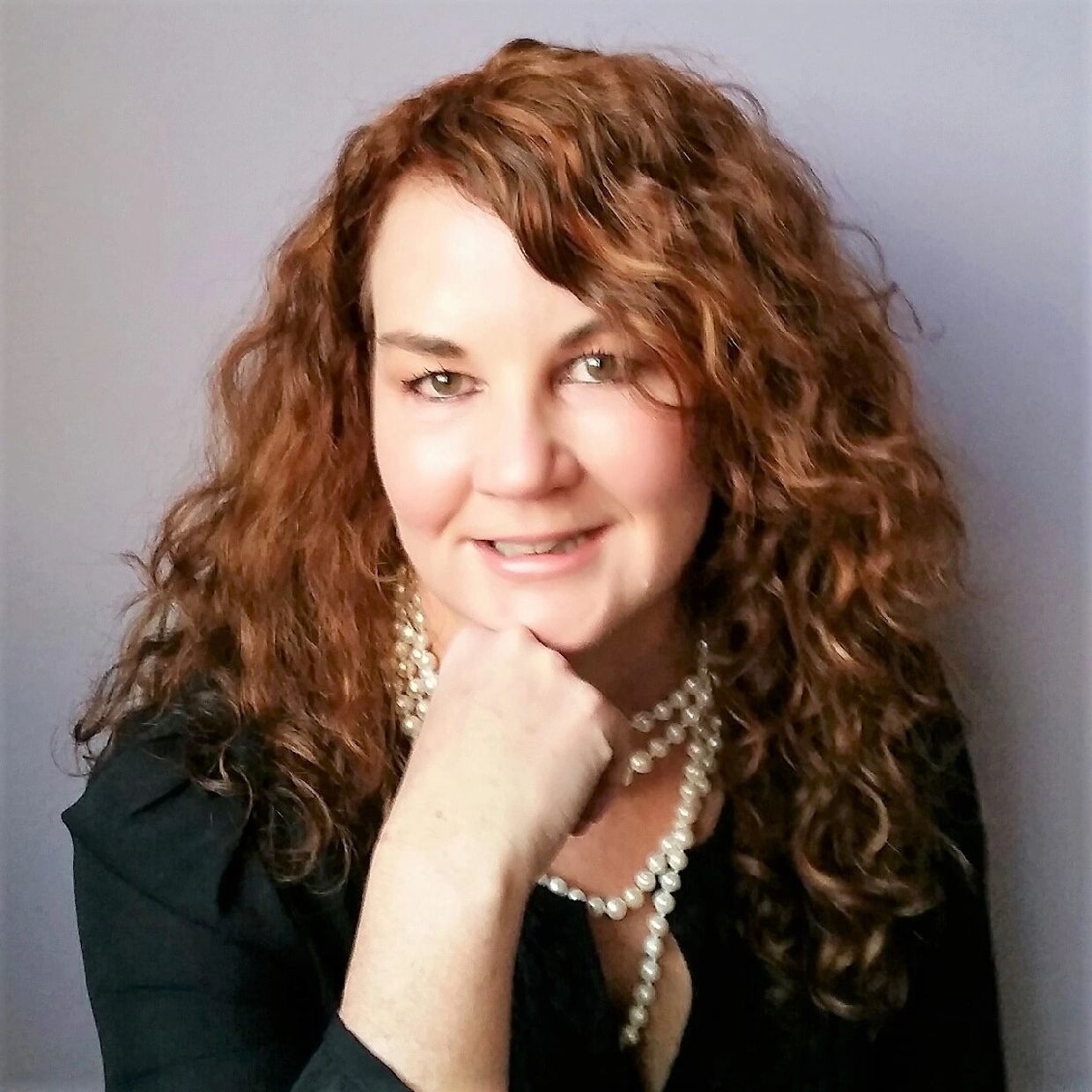 Smiling woman with curly red hair wearing a black top and pearl necklace.