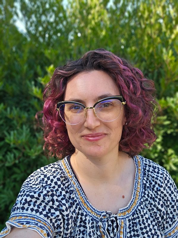 A white woman with glasses and curly burgundy hair against a green backdrop.