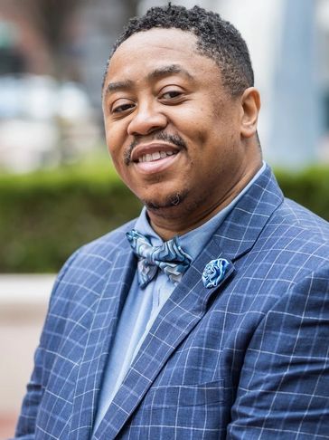 Smiling man in a blue checkered suit with a bow tie and lapel flower.