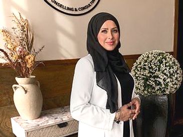 Woman in a white blazer and black hijab smiling indoors.