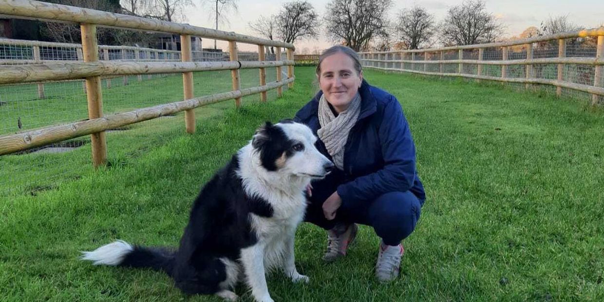 A woman crouches beside a black and white dog in a grassy fenced area.