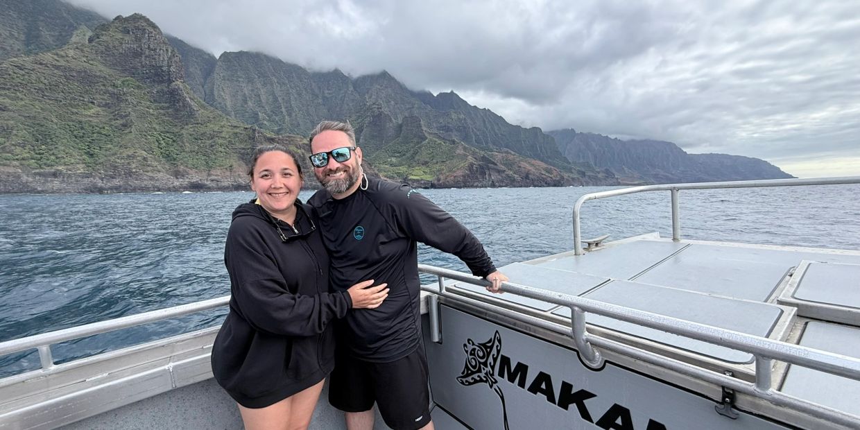 Couple smiling on a boat with mountains and ocean in the background.
