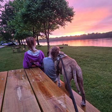 Two girls and a dog watch a colorful sunset by the river from a wooden picnic table.