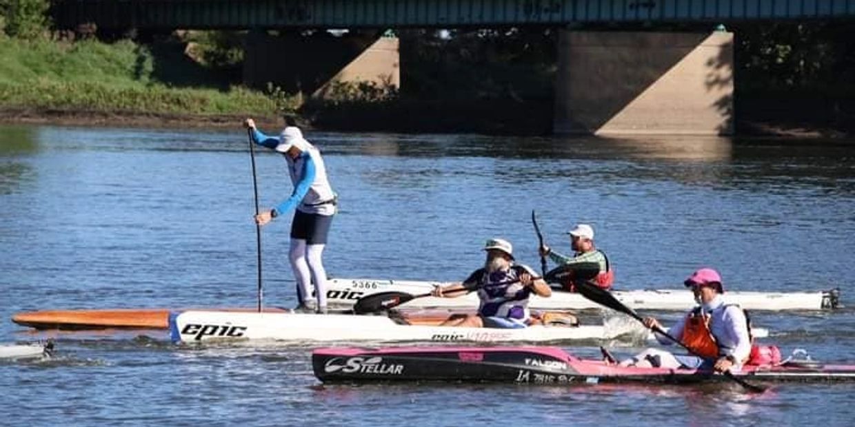 Three people paddle on kayaks and a stand-up paddleboard in a river near a bridge.