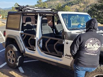 Person standing near a white Jeep with no doors in a mountainous area.