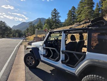 A silver Jeep with open doors parked by a scenic mountain road.