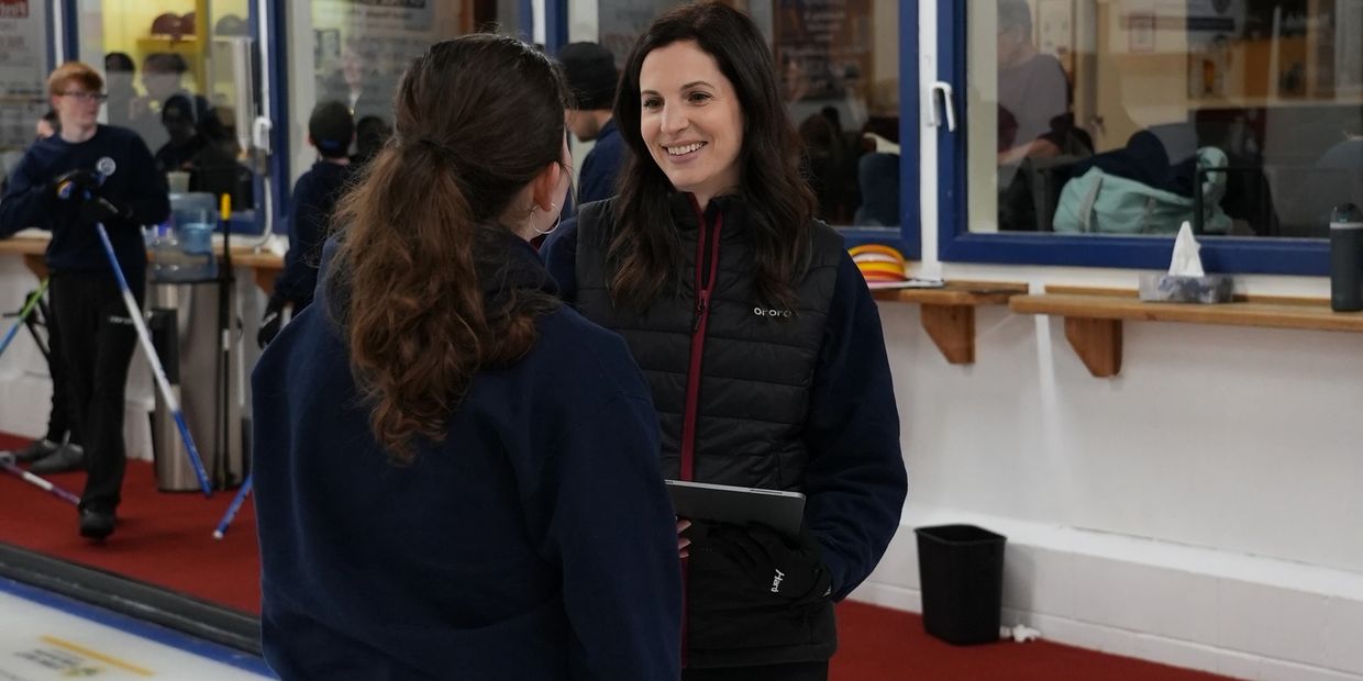 Two women chatting indoors near a curling rink with players in the background.