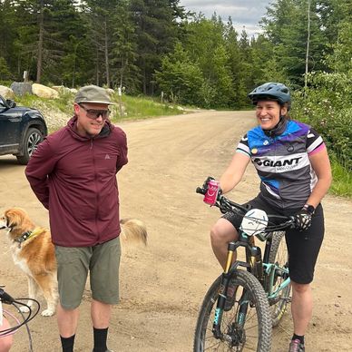 Two smiling friends with a dog and bikes on a forest dirt road.