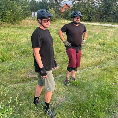 Two men in helmets standing on a grassy field with trees and a cabin in the background.