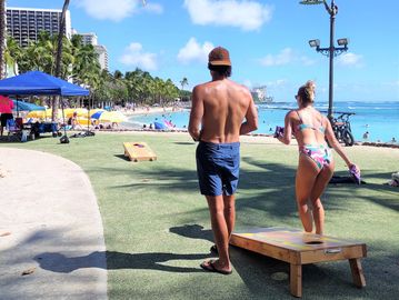 Couple playing Cornhole with palm trees and beach in the background, Waikiki, Hawaii.