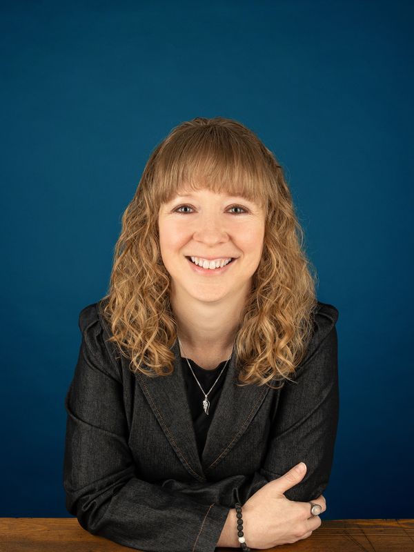 Smiling woman with curly hair in a black blazer against a blue background.